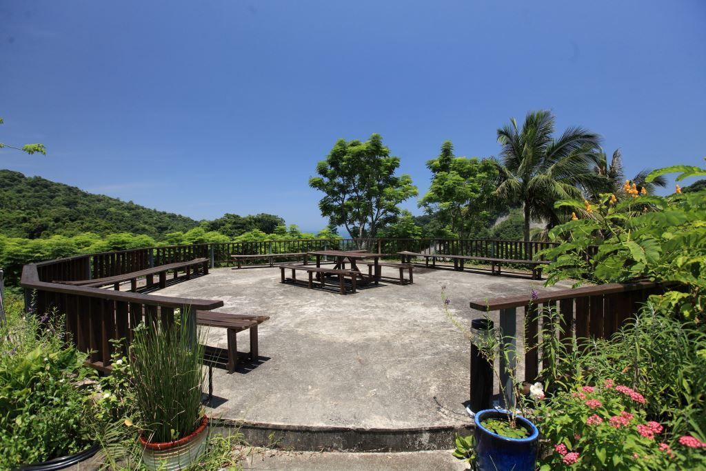 An outdoor seating area with picnic tables and benches, surrounded by lush greenery and a clear blue sky.