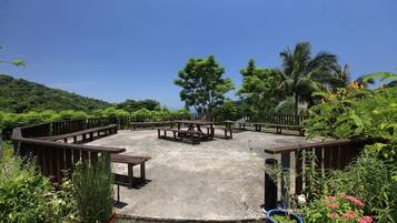 An outdoor seating area with picnic tables and benches, surrounded by lush greenery and a clear blue sky.