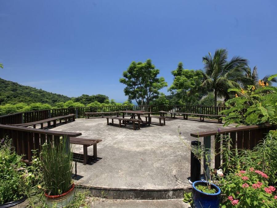 An outdoor seating area with picnic tables and benches, surrounded by lush greenery and a clear blue sky.