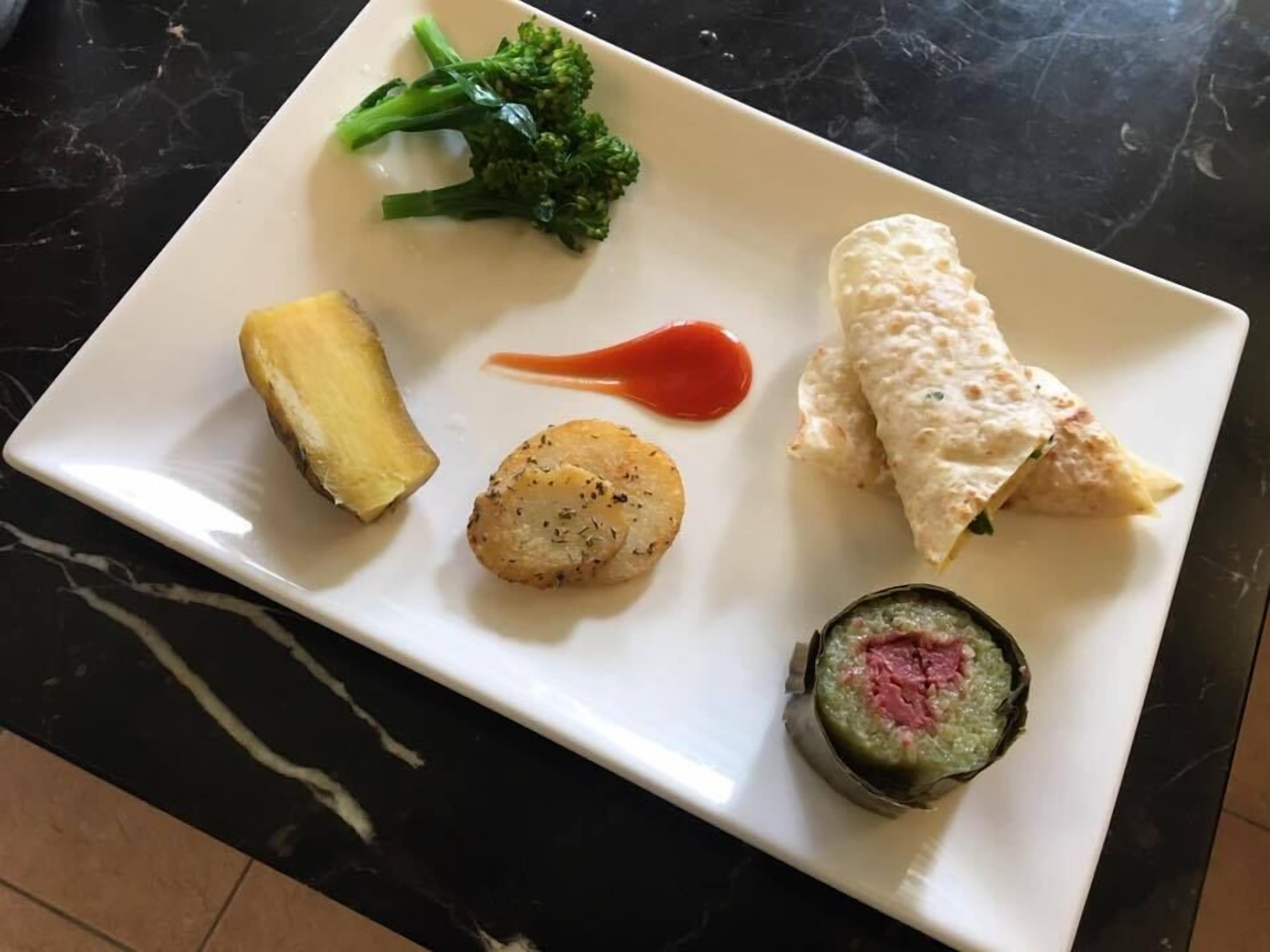 A white rectangular plate with various food items including broccoli, a rolled omelette, a scallop, a piece of ginger, a red sauce, and a sushi roll.
