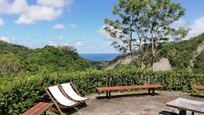 A scenic outdoor area with a view of the ocean, surrounded by lush greenery and equipped with wooden benches and lounge chairs. - Juhu Farmstay (Changbin)