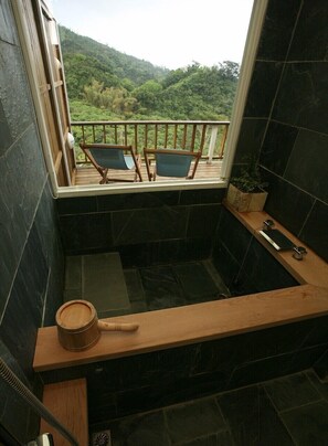 Un baño con una bañera de madera, un ventanal con vistas a un bosque y dos sillas en un balcón.