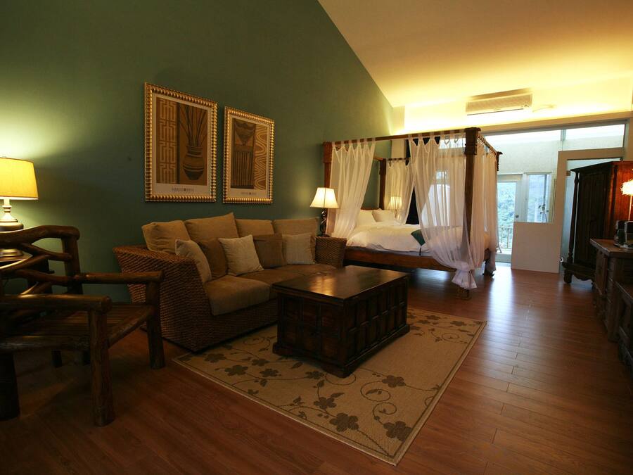Una sala acogedora con sofá, sillón y mesa de centro. Se observan dos cuadros en la pared, una cama con dosel y un escritorio con lámpara.