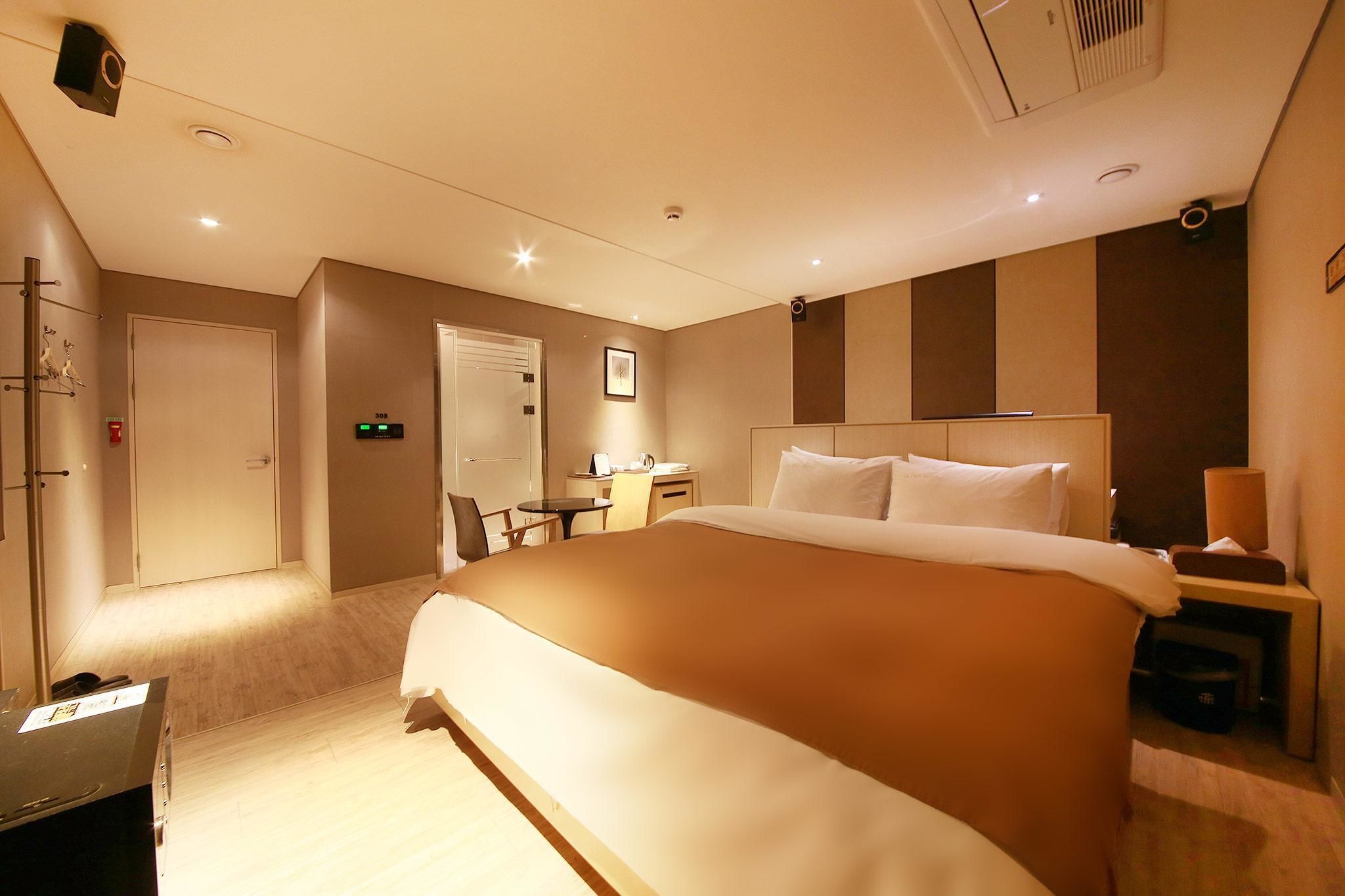 A hotel room with a large bed, a desk, and a chair. There is a door, a wall-mounted air conditioner, and a small table with a lamp.