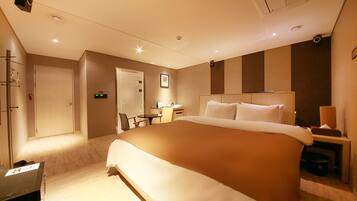 A hotel room with a large bed, a desk, and a chair. There is a door, a wall-mounted air conditioner, and a small table with a lamp.