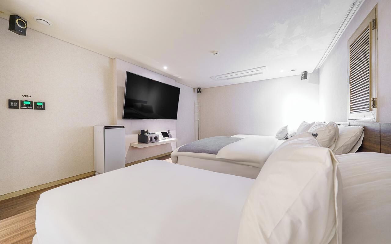 A hotel room with two beds, a flat-screen TV, a desk with a radio, and a window with blinds.