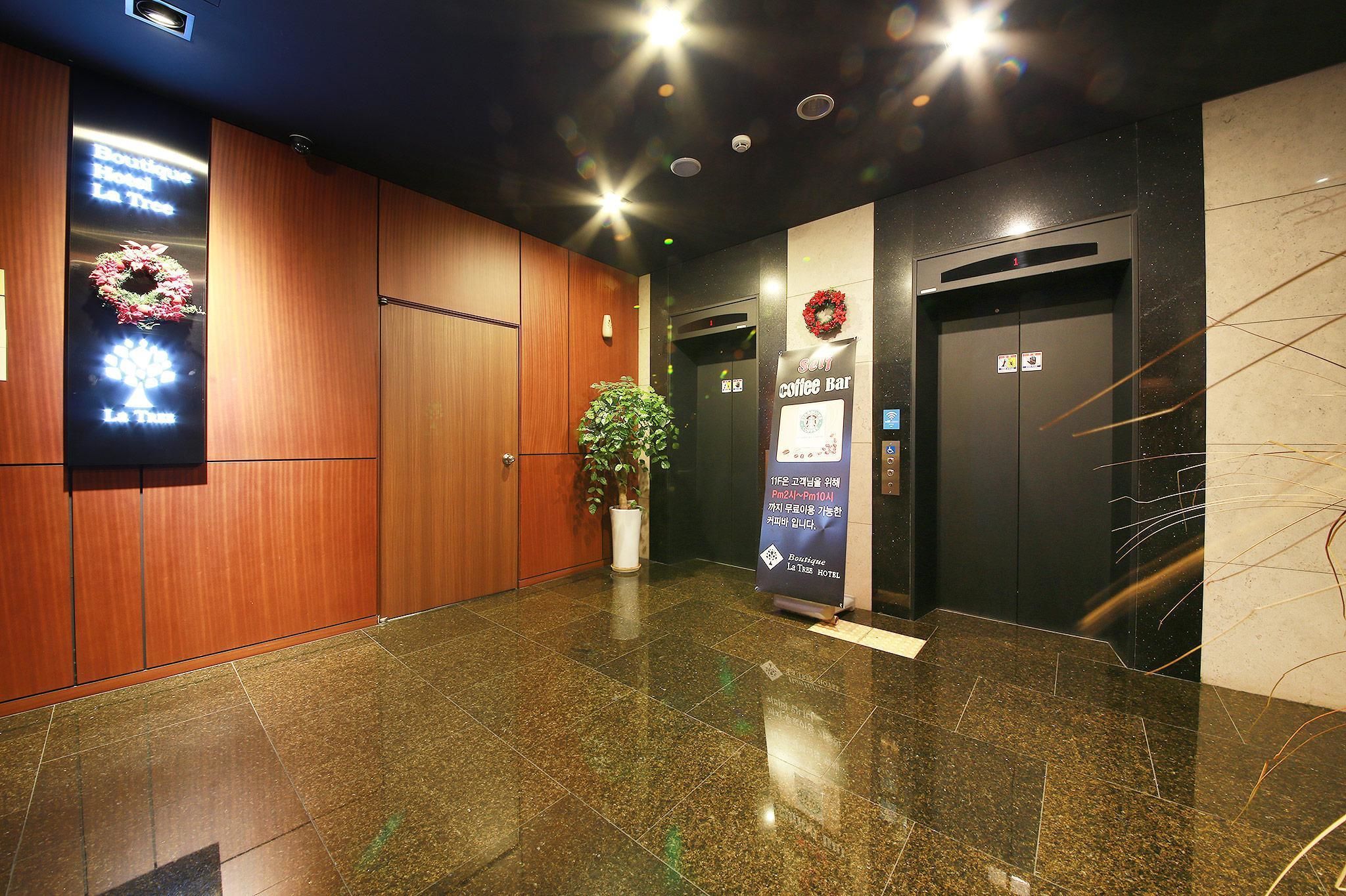 A hotel lobby with a wooden reception desk, a potted plant, a sign for a coffee bar, and an elevator.