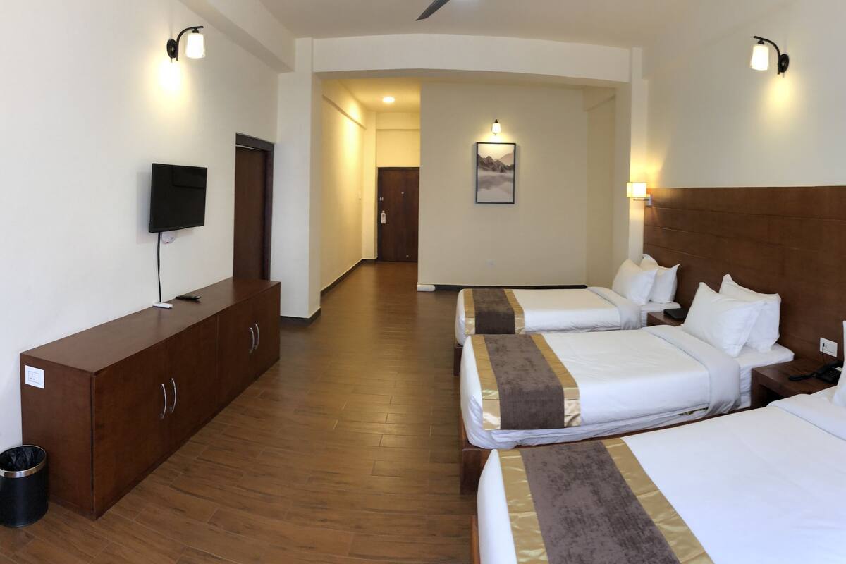 A hotel room with three single beds, a wooden wardrobe, a television, and a hallway leading to another room.