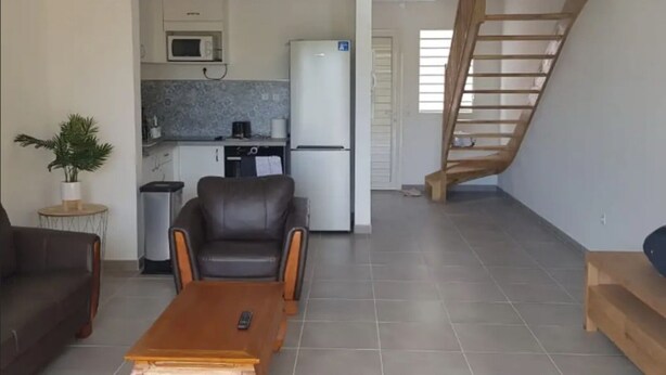 Ein Wohnzimmer mit einer Couch, einem Couchtisch und einer Küchenzeile inklusive Mikrowelle, Kühlschrank und einer Treppe.