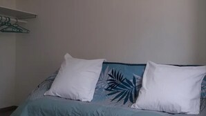 Ein Bett mit einer blau-weißen blumigen Tagesdecke, zwei weiße Kissen und ein grüner Kleiderbügel an einer weißen Wand.