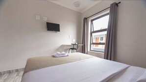 A hotel room with a bed, a desk, a chair, a TV mounted on the wall, and a window with curtains. - Ecomotel Germiston (Germiston)