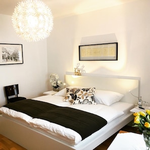 Un lit bien fait, agrémenté d’un coussin à motifs noir et blanc, d’une tête de lit blanche et d’une étagère murale ornée d’objets décoratifs.
