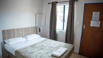 A hotel room with a bed, a window with curtains, a door, and a wall-mounted light.