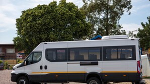 A white van parked on a gravel surface with a blue roof rack, surrounded by trees and buildings. - Ecomotel O.R Tambo Intl (Kempton Park)