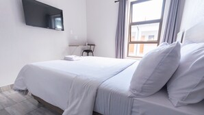 A hotel room with a bed, a flat-screen TV mounted on the wall, a desk with a chair, and a window with curtains. - Ecomotel O.R Tambo Intl (Kempton Park)