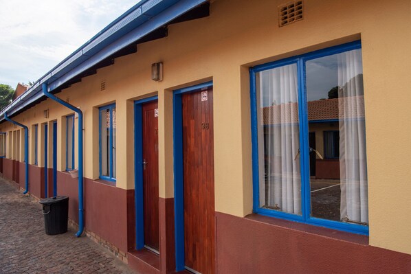 A row of hotel rooms with wooden doors and blue trim, a trash bin, and a brick-paved walkway.