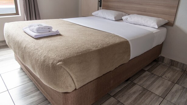 A neatly made bed with a beige blanket and white pillows, a wooden headboard, and a window with curtains. - Ecomotel O.R Tambo Intl (Kempton Park)