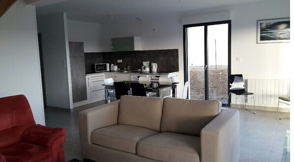 A modern living room with a beige sofa, a red armchair, and a kitchen area with a dining table and chairs. - New house near the beaches of the landing (Saint-Côme-de-Fresné)
