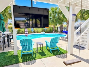 A pool area with a TV screen, two chairs, a small table, and a swing.