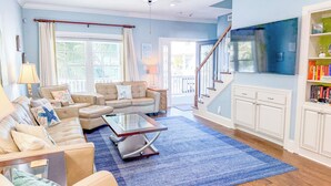 A spacious living room with a beige sofa set, a coffee table, and a blue rug. There is a staircase leading to an upper level, and a bookshelf filled with books. - Luxury 4Bedroom/ 4 Bath with Private Salt-Water Pool! (North Myrtle Beach)