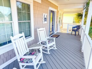 A covered porch with two rocking chairs and a table, featuring a brick wall and large windows.