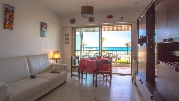 A living room with a white sofa, a small table with a red tablecloth, and a balcony with a view of the sea. - Seafront studio with terrace, near beaches and center, Free Wifi (Menton)