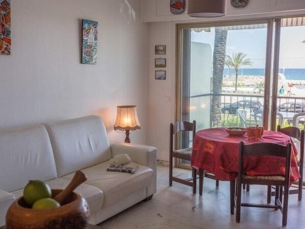 A living room with a white sofa, a table with a red tablecloth, and a view of the beach through large windows. - Seafront studio with terrace, near beaches and center, Free Wifi (Menton)