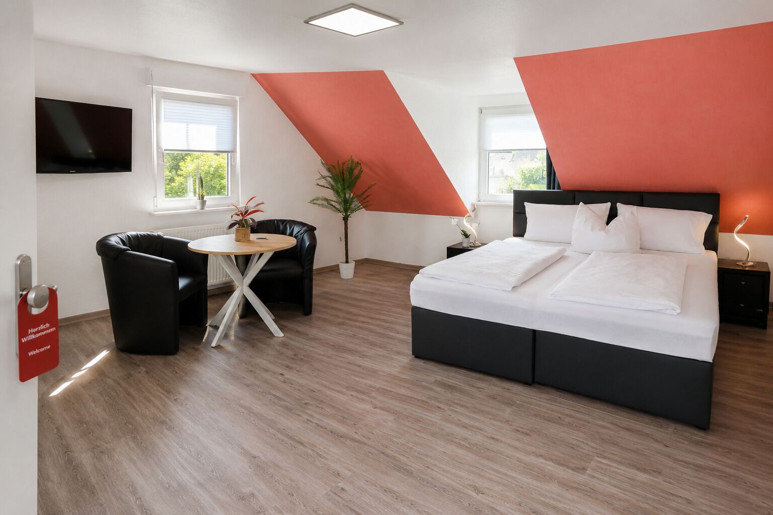 A modern hotel room with a bed, a desk, and a chair. There is a TV mounted on the wall, a window, and a red accent wall.