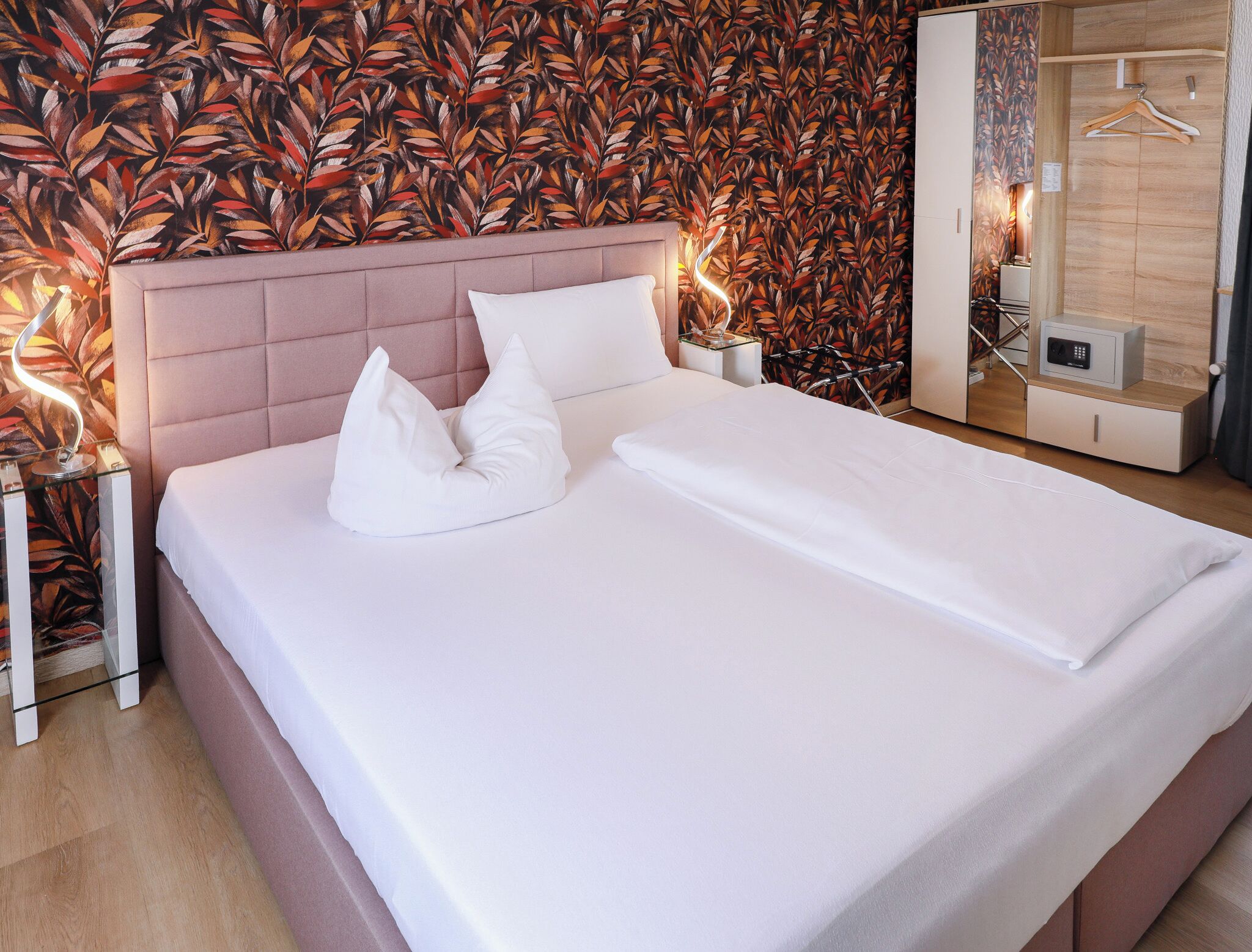 Una cama bien hecha con sábanas y almohadas blancas, un cabecero tapizado en rosa, y una pared con un diseño de hojas.
