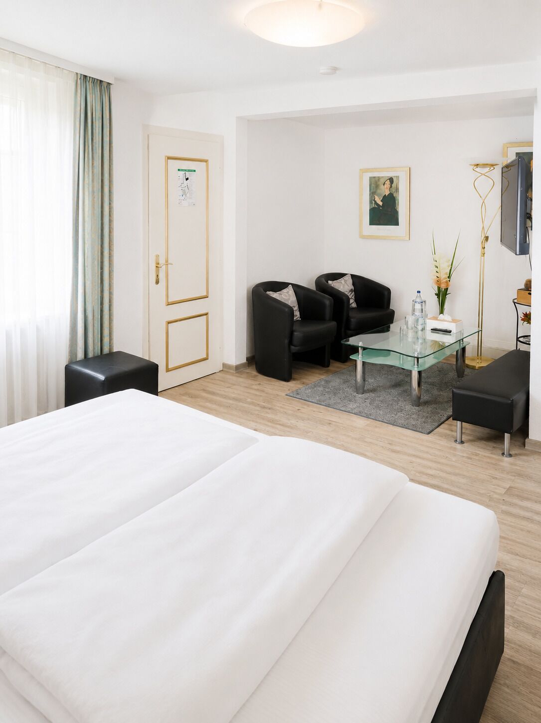 A hotel room with a large bed, two black armchairs, a glass coffee table, and a desk with a flat-screen TV.