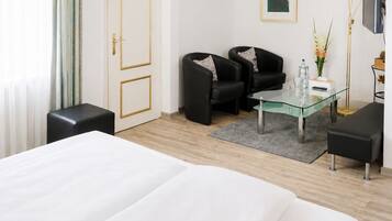 A hotel room with a large bed, two black armchairs, a glass coffee table, and a desk with a flat-screen TV.