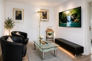 Una sala de estar moderna con una mesa de centro de vidrio, sofás de cuero, un banco, una lámpara de pie y un televisor grande montado en la pared.