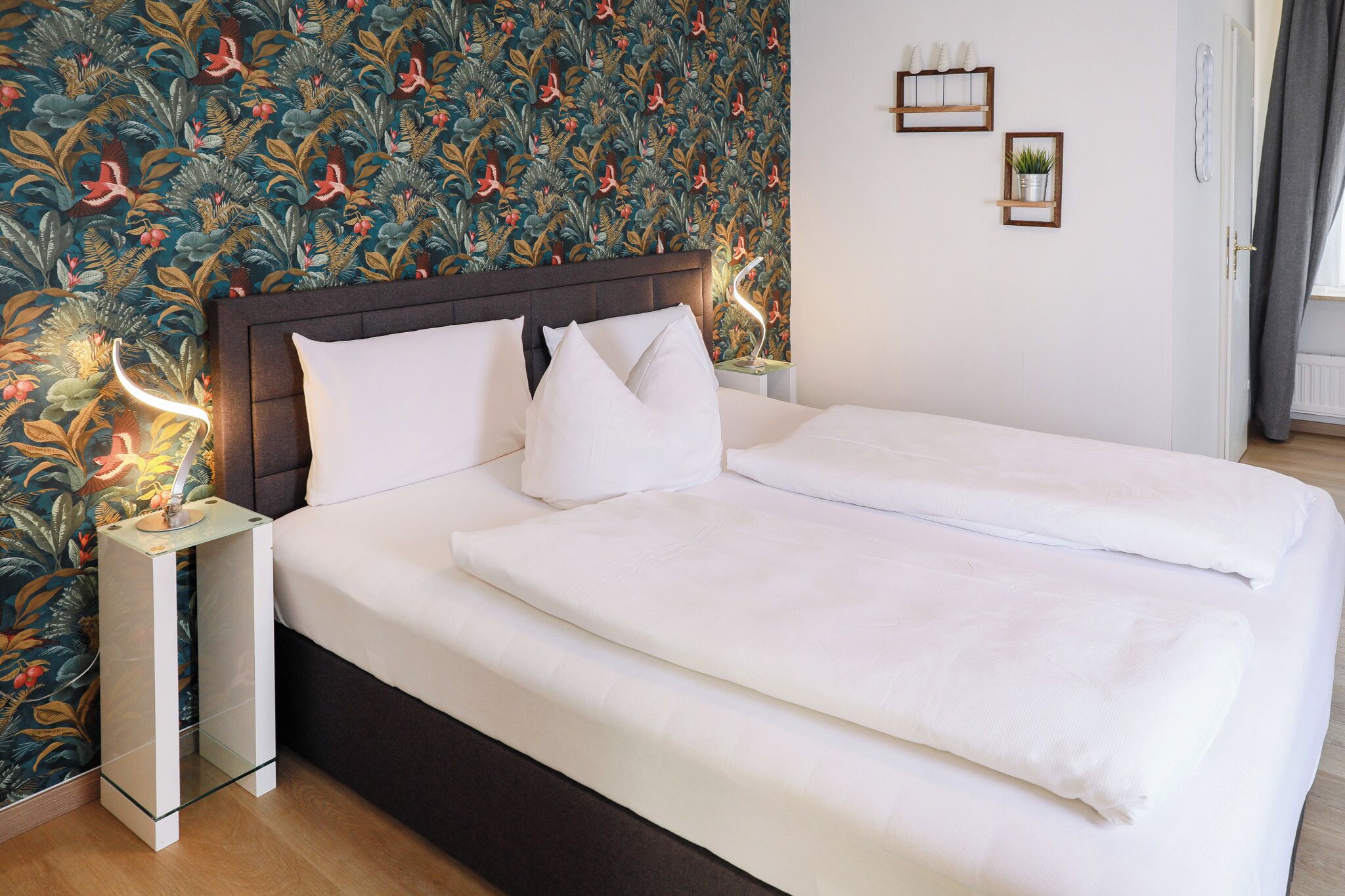 Quarto de hotel com uma cama grande, mesa de cabeceira com abajur, papel de parede floral e uma pequena planta na parede.