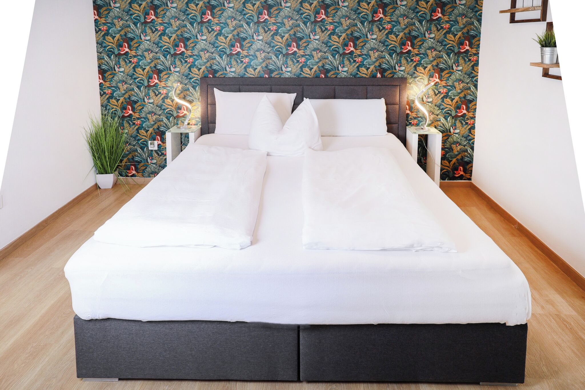 A bed with a dark headboard, white bedding, and a floral wallpaper background.