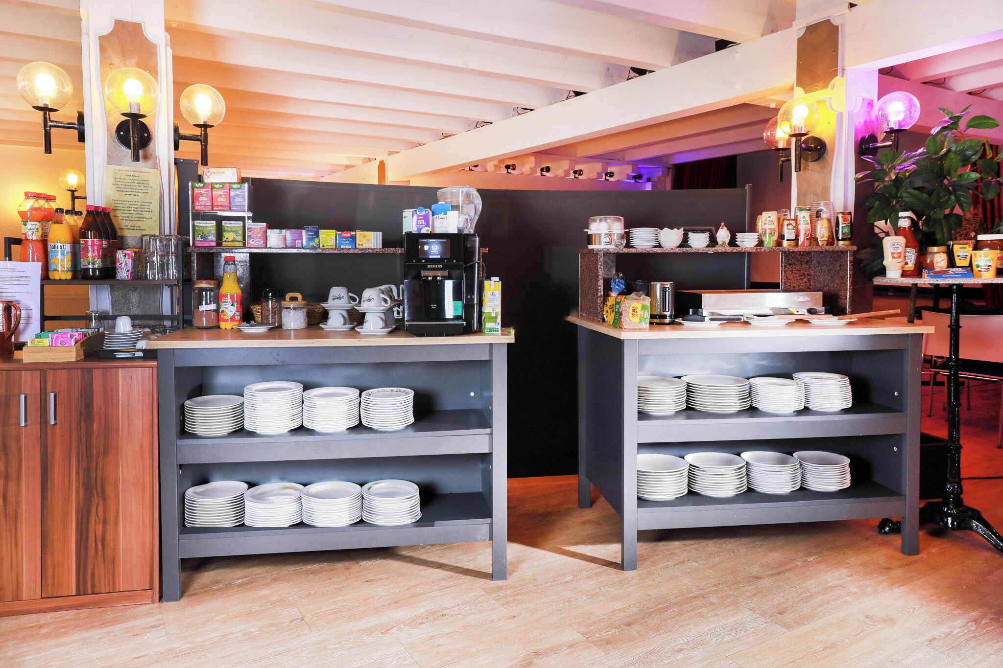 Un área de desayuno con varias máquinas de café, una variedad de alimentos para el desayuno y platos apilados ordenadamente en estantes.