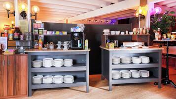 Un área de desayuno con varias máquinas de café, una variedad de alimentos para el desayuno y platos apilados ordenadamente en estantes.