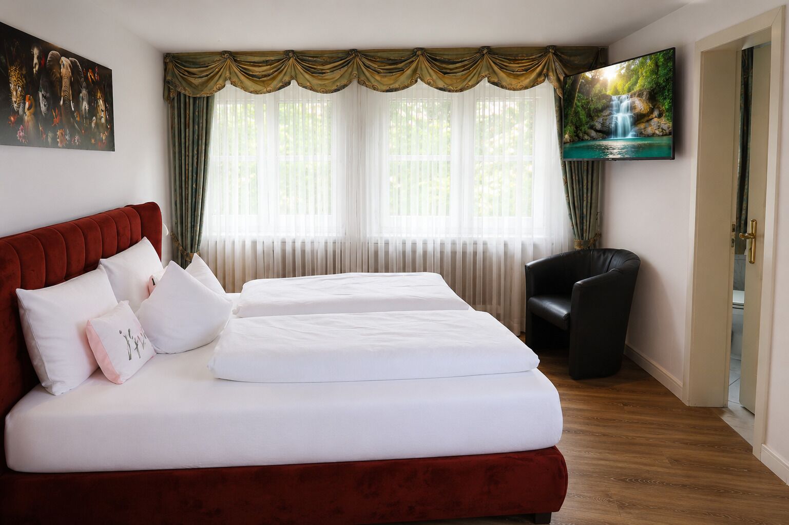 Habitación de hotel con cama doble, cabecera roja, televisor montado en la pared y una ventana con cortinas traslúcidas.