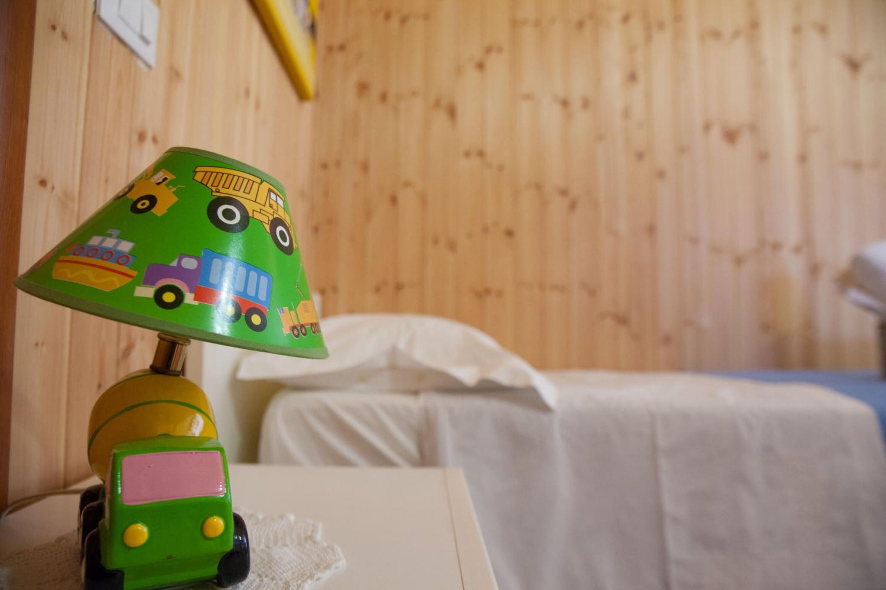 En säng med ett vitt lakan, en grön lampa med en tecknad design och en liten leksaksbil på nattduksbordet.