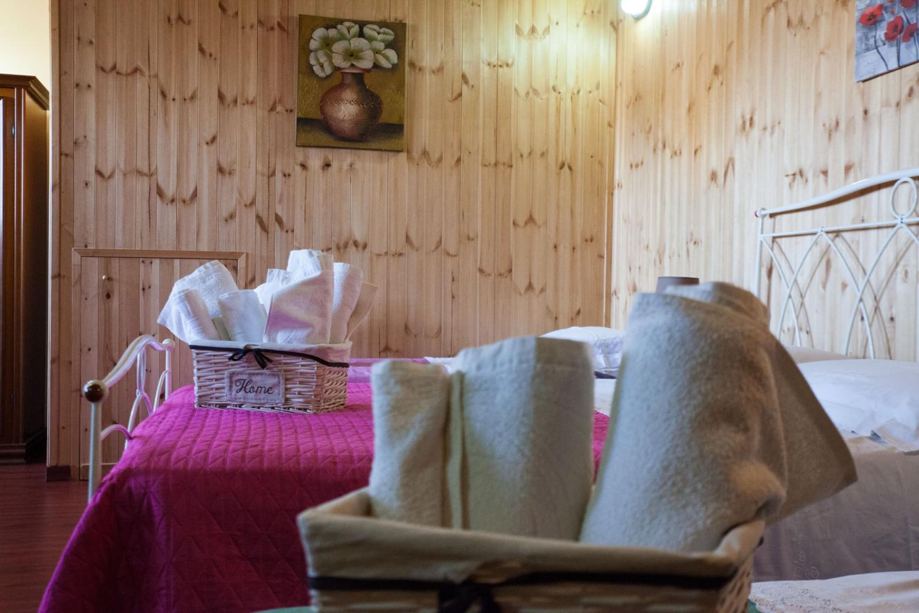 Een kamer met een houten wand, een bed met een wit metalen hoofdeinde, een roze dekbedovertrek, een rieten mandje met de tekst 'Thuis' en een schilderij van bloemen.