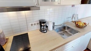 Une cuisine avec des meubles blancs, une plaque de cuisson à induction noire, une machine à café, un évier et un panier de fruits.