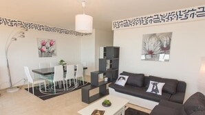 Ein modernes Wohnzimmer mit einer schwarzen Couch, einem weißen Couchtisch und einem Glastisch mit Stühlen. Es gibt ein Bücherregal und eine Stehlampe.