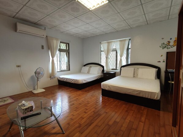 Une chambre d’hôtel avec deux lits, un ventilateur sur pied, une petite table et une fenêtre avec des rideaux.