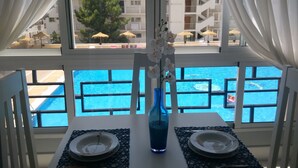 Una mesa de comedor con dos platos, un jarrón azul con flores blancas y vista a una piscina a través de una ventana con cortinas blancas.