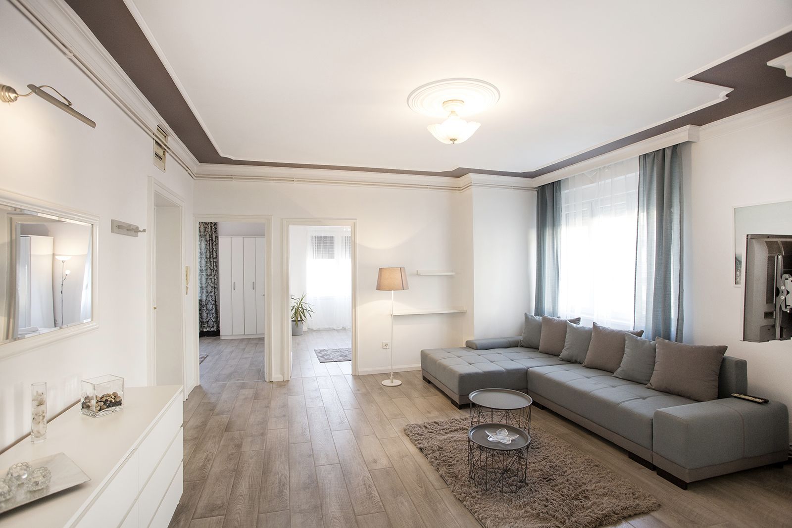 Un salon moderne avec un canapé d’angle gris, une table basse ronde et un tapis moelleux. Un couloir mène à une autre pièce, où se trouvent une porte et une plante.