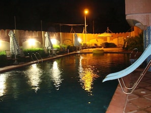 Une piscine nocturne, dotée de chaises longues et de parasols, entourée d’un petit muret et d’un lampadaire.