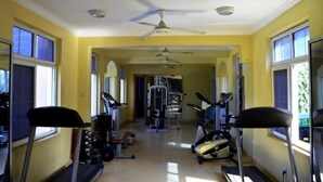A gym with treadmills, exercise bikes, and weight machines, with large windows and a ceiling fan. - Abla Apartment (Dar es Salaam)