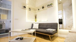Ein modernes Wohnzimmer mit einem grauen Sofa, einem weißen Couchtisch und einem kleinen, weißen Beistelltisch mit einer Fernbedienung. An der weißen Wand befinden sich eine Uhr und Regale mit Pflanzen.