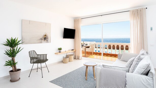 A modern living room with a large balcony overlooking the sea, a flat-screen TV, and a minimalist decor.