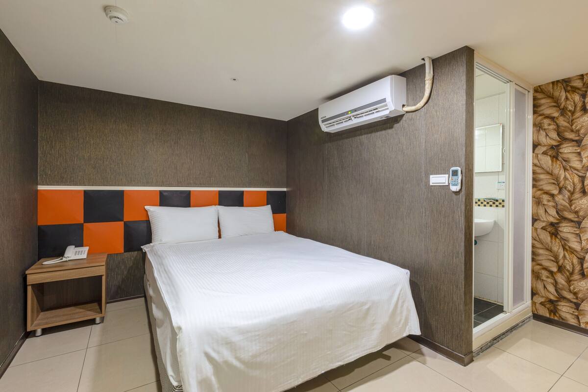 A hotel room with a bed, a nightstand with a phone, an air conditioning unit, and a shower area.