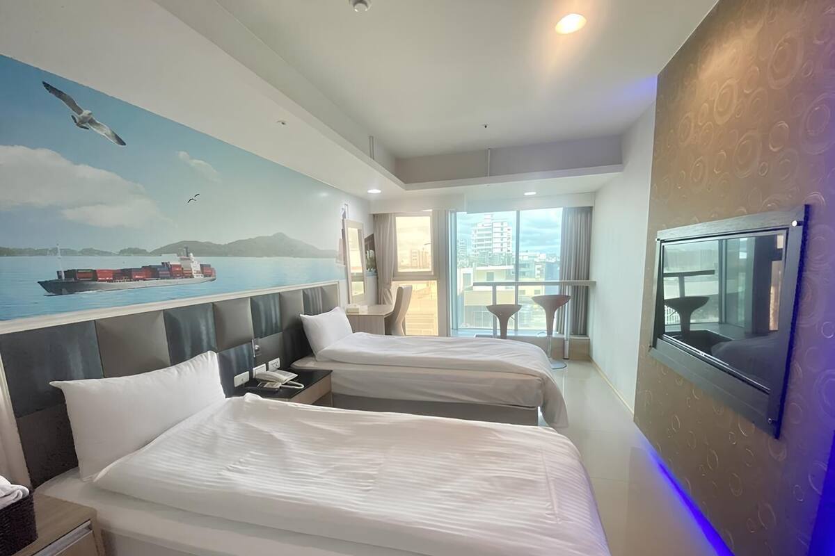 A hotel room with two beds, a large window with a view of the city, a painting of a ship on the wall, and a TV mounted on the wall.